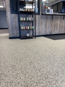 A professional photo of the interior of a veterinary office or kennel. The floor is covered with a durable, light-colored epoxy coating with a speckled pattern of black, gray, and brown flakes. A small black shelf unit is in the middle of the room, holding several jars of treats. The walls behind the counter are covered with wood-like paneling. A small paw print is visible on the bottom shelf, and a blue "BBB" logo is on a sticker next to the counter.