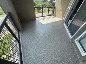
A photo of a residential balcony or patio with a newly installed epoxy floor coating. The floor is light gray with black, white, and gray flakes, and has a glossy finish. The walls are a light brown stucco. A set of sliding glass doors with a white frame is on the right, and a metal railing overlooks a green, outdoor area with trees and hills in the distance. The sun is shining brightly, casting shadows on the floor.