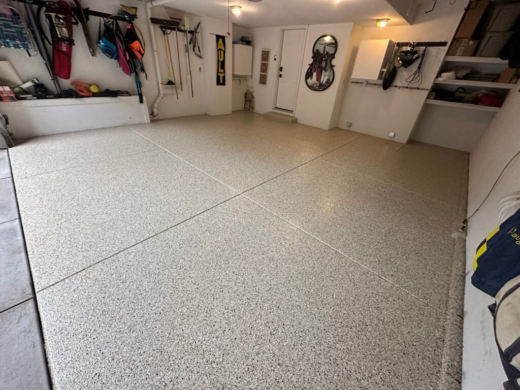 A professional photo of the interior of a clean, organized garage. The floor is covered with a new, light-colored polyurea coating with speckled brown and gray flakes. The floor has a slight sheen and is marked by four clean lines where the coating meets. The walls are white, and various sports equipment, tools, and storage racks are neatly hung or stored. The far wall has a white door.