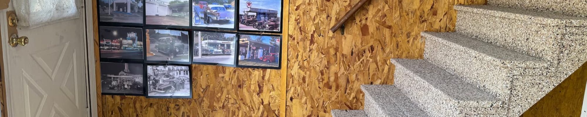 A photo of an indoor space that appears to be a basement or a garage. The floor is covered with a new, light-colored epoxy coating with black and brown flakes. A staircase with the same speckled coating leads up from the floor, with a dark wooden handrail. The walls are made of particle board, with a black circular clock and a large grid of framed photos of cars and other vehicles on display. A white door with a window is visible on the far left.